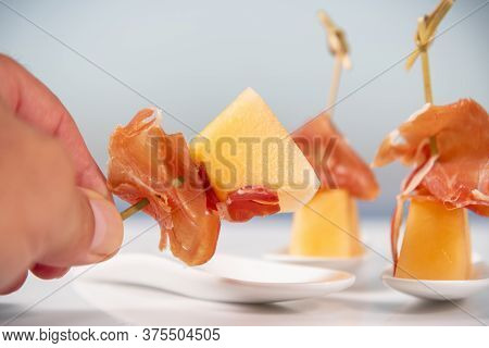 A Male Hand Holding A Skewer Made Of A Small Melon Piece And A Thin Slice Of Cured Ham,