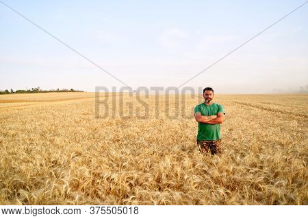 Happy Farmer Proudly Standing In Wheat Field With Arms Crossed On Chest. Agronomist Wearing Corporat