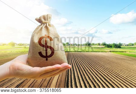 A Hand Holds Out A Dollar Money Bag On A Background Of A Farm Field. Lending Farmers And Agricultura
