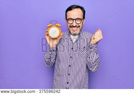 Middle age man wearing glasses holding vintage alarm clock over isolated purple background screaming proud, celebrating victory and success very excited with raised arm