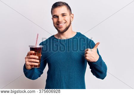 Young handsome man dirnking cola refreshment beverage smiling happy and positive, thumb up doing excellent and approval sign 