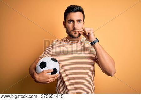 Handsome player man with beard playing soccer holding footballl ball over yellow background mouth and lips shut as zip with fingers. Secret and silent, taboo talking