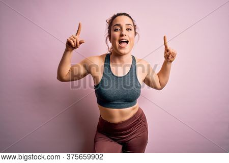 Young beautiful blonde sportswoman doing sport wearing sportswear over pink background smiling amazed and surprised and pointing up with fingers and raised arms.