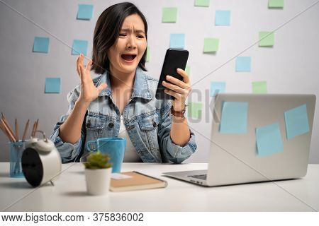 Asian Woman Angry Looking At Smart Phone Reading News And Shouting At Home Office. Wfh. Work From Ho