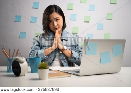 Asian Woman Holding Hands In Prayer Looking At Camera And Working On A Laptop At Home Office. Wfh. W