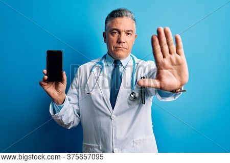 Middle age handsome grey-haired doctor man holding smartphone showing screen with open hand doing stop sign with serious and confident expression, defense gesture