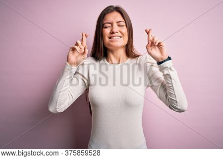 Young beautiful woman with blue eyes wearing casual white t-shirt over pink background gesturing finger crossed smiling with hope and eyes closed. Luck and superstitious concept.