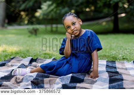 Portrait Of Little Cute Happy African Girl In Blue Dress, Sitting On Checkered Blanket In Summer Par