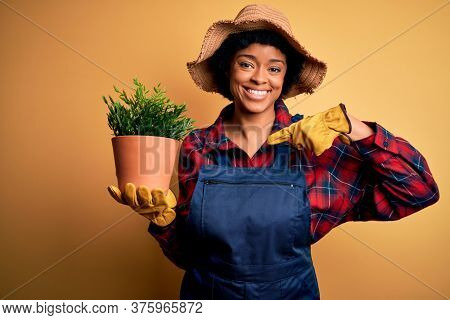 Young African American farmer woman with curly hair wearing apron holding pot with plants with surprise face pointing finger to himself