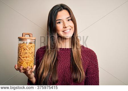 Young beautiful girl holding bottle of dry Italian pasta macaroni over white background with a happy face standing and smiling with a confident smile showing teeth