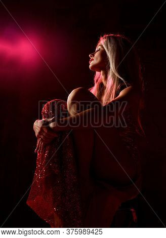 Young Beautiful Blonde Woman In Sexy Red Shiny Dress With Open Legs Sitting On Chair, Holding Tight 