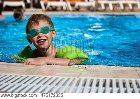 Little Boy Swims Independently In A Large Pool In Special Glasses And Sleeves Of Green Color. Indepe