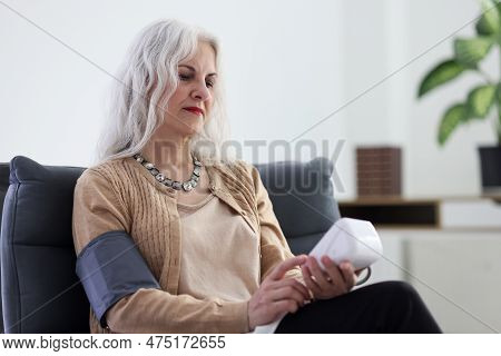Senior Woman Uses Digital Tonometer To Measure Blood Pressure Sitting In Armchair At Home. Contempor