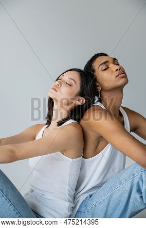 Young And Trendy Interracial Couple In White Tank Tops Sitting Back To Back On Grey Background.