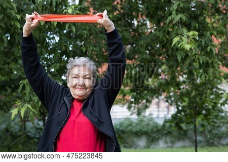 Smiling Elderly Woman Is Doing Exercises With Fitness Elastic Band Outdoors In The Yard. Active Life