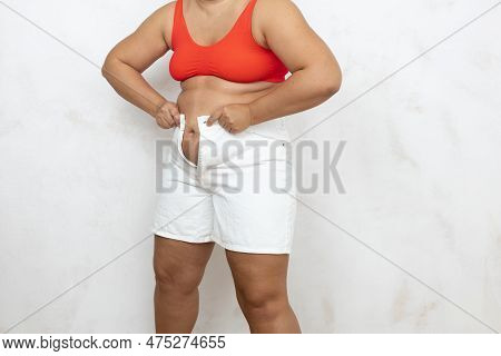 Hands Of Overweight Woman Pull White Denim Shorts Over Large Belly, White Background. Woman In Red U
