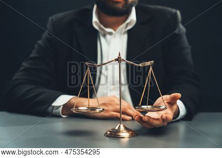 Lawyer In A Black Suit Sits Attentively On His Office Desk With A Golden Scale Balance, Symbol Of Le