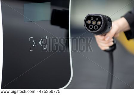 Focus Closeup Hand Holding Ev Plug For Electric Vehicle Pointing At Camera With Charging Station At 