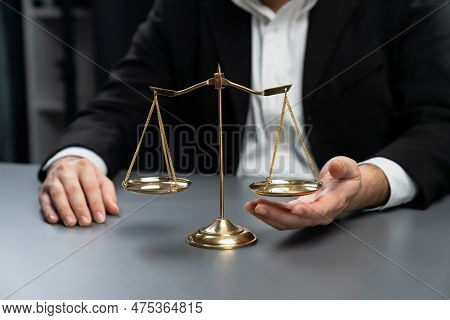 Lawyer In A Black Suit Sits Attentively On His Office Desk With A Golden Scale Balance, Symbol Of Le