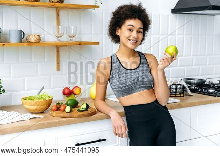 Lovely Positive Confident African American Girl In Sportswear Stand In The Kitchen, Holds An Apple I
