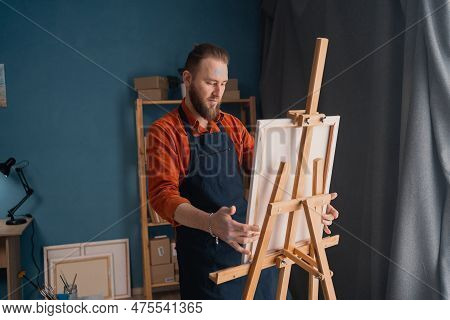 Portrait Caucasian Bearded Male Artist In An Apron In A Home Studio Stands Near An Easel Holds A Can