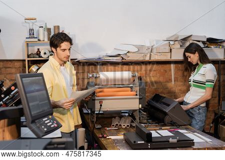 Good Looking Typographer Looking At Printed Paper Near Pretty Colleague.