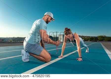 Instructor And Athlete Runner On The Track. The Athletics Marks The Time In The Plank Exercise On A 