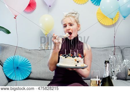 Sad, Upset Woman In Cocktail Dress And Party Cap Eating Festive Cake While Sitting On The Sofa And C
