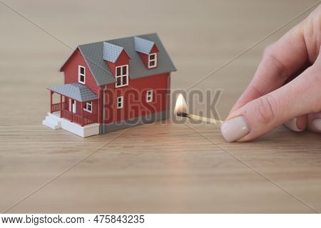 Hand Of Woman Holding Match Ready To Burn House Standing On Wooden Table. Small Model Of Two-story T