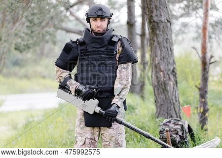 A Man In A Military Uniform And Bulletproof Vest Works In The Forest With A Metal Detector. A Minesw