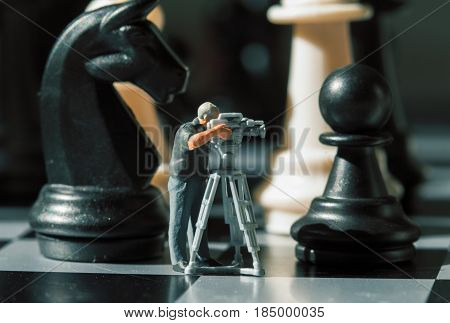 Cameraman miniature doll and chess figures on board. Filming the checkmate game. Black knight and pawn in camera. News making about chess. Videographer macro photo. Making movie with chess figures