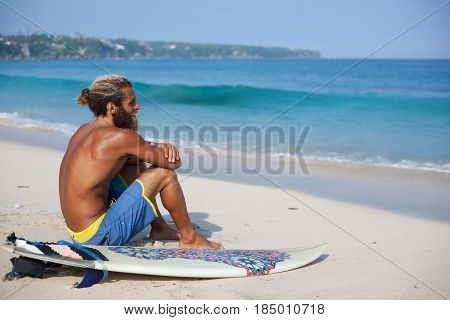 Attractive bearded curly man with a surfboard is seating with his back on a sand and looking to the ocean