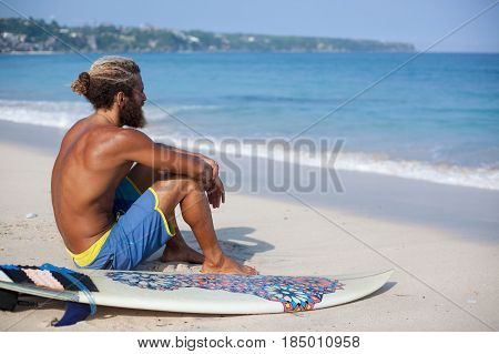Horizontal portrait of an attractive bearded curly man with a surfboard is seating with his back on a sand and looking to the ocean