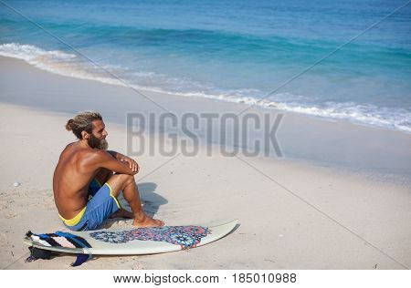 Horizontal portrait of an attractive bearded curly man with a surfboard is seating with his back on a sand and looking to the ocean located on the left side of the frame