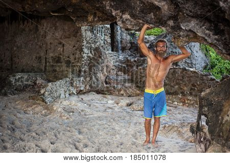 Full-length portrait of an attractive bearded curly man is standing with his hands up on a sand under a rock and looking to the camera