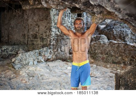 Attractive bearded curly man is standing with his hands up on a sand under a rock and looking to the camera