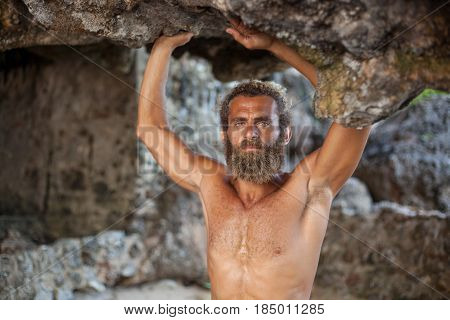 Half-length portrait of an attractive bearded curly man is standing with his hands up on a sand under a rock and looking to the camera