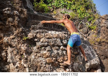 Full-length horizontal portrait of a handsome bearded curly man with his back is swarming up a rock