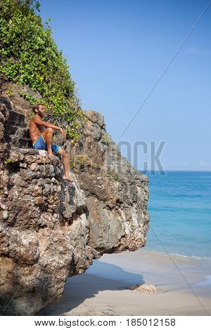 Vertical portrait of an attractive bearded man sitting on a rock close the ocean located on the left side of the frame