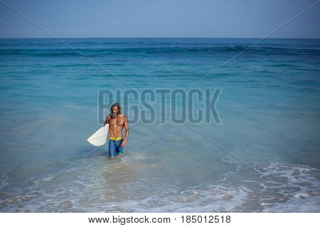 Attractive bearded curly wet man in blue shorts and with naked torso with white surfboard coming out of the ocean located on the left side of the frame