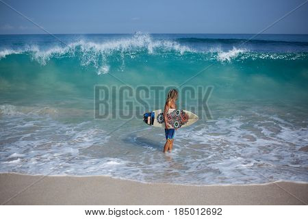 Handsome muscular curly blond man with a surfboard standing on the shore of the sea and looking at the big wave rising behind him