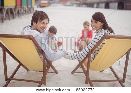 Portrait of smiling parents relaxing on launge chairs with children playing in background at beach