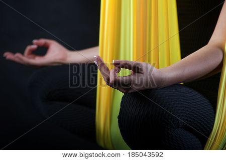 Cropped shot of young woman sitting in yellow hammock and meditating on black background. Girl practicing fly yoga, studio shot. Body balance and antigravity exercises. Wellness and bodycare concept