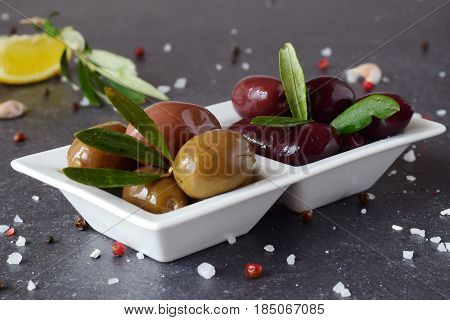 Black and green olives in a white plate on a grey abstract background with lemon, garlic, spices. Mediterranean food. Healthy eating concept
