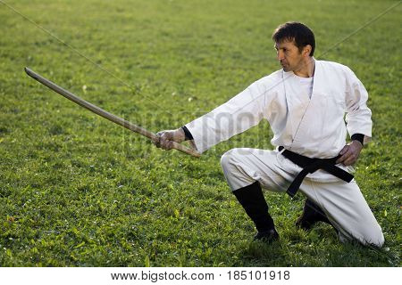 Man in white kimono with wooden sword is practicing martial arts on the grass outdoors.
