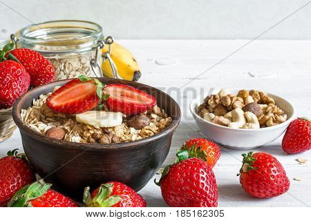 bowl of oat muesli with strawberry, banana, granola and nuts with ingredients for healthy breakfast on white wooden table. close up