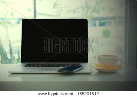 Laptop blank black screen copy space and smartphone on table with coffee cup and window background (vintage tone)