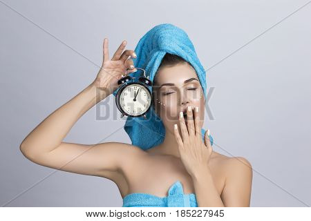 Tired yawning woman in towel on head and creme on face holding alarm clock showing midnight night creme skincare concept