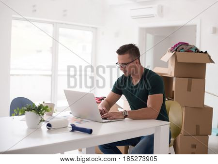 Young man moving in a new home. Man at the table using notebook laptop computer and plans with boxes around him