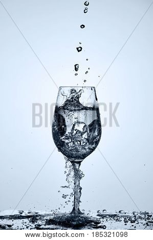 Drinking transparent water is poured into a wineglass standing on the glass with water splashing against light background. Useful and natural product. Care for the environment and ecology. Concept of the healthy lifestyle.